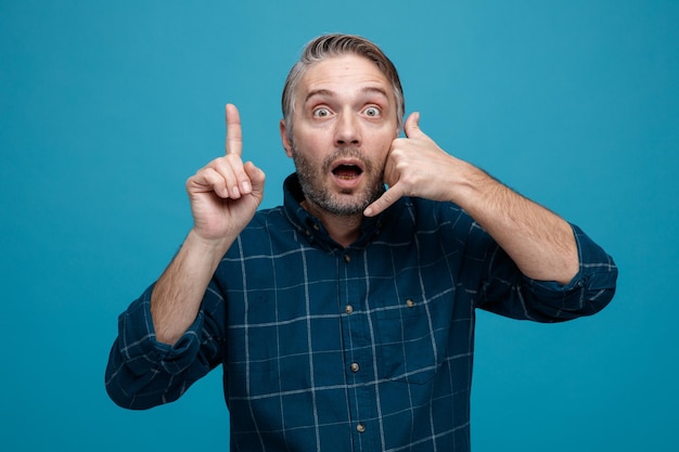 Middle age man with grey hair in dark color shirt looking at camera being surprised making call me gesture with hand pointing with index finger up standing over blue background