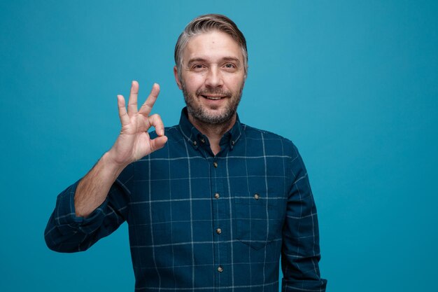 Middle age man with grey hair in dark color shirt looking at camera happy and positive smiling showing ok sign standing over blue background