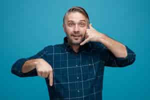 Free photo middle age man with grey hair in dark color shirt looking at camera making call me gesture with hand pointing with index finger down smiling standing over blue background