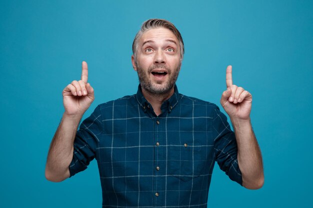 middle age man with grey hair in dark color shirt looking up happy and surprised pointing with index fingers up standing over blue background