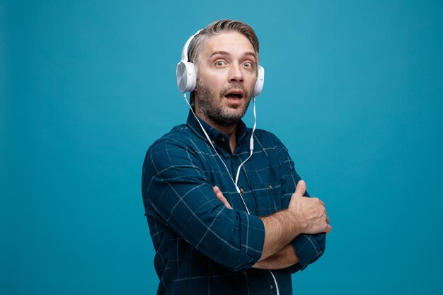 Middle age man with grey hair in dark color shirt with headphones looking at camera amazed and surprised with arms crossed on his chest standing over blue background