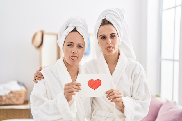 Free photo middle age woman and daughter wearing bath robe holding heart card sticking tongue out happy with funny expression.