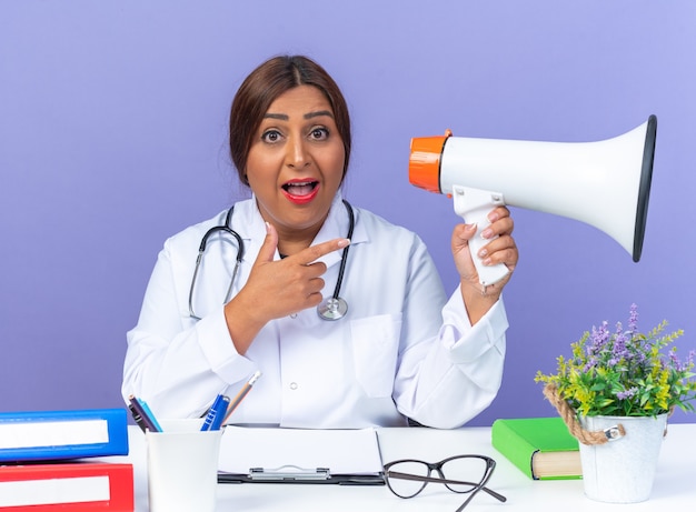 Middle age woman doctor in white coat with stethoscope holding megaphone pointing with index finger at it happy and surprised sitting at the table on blue