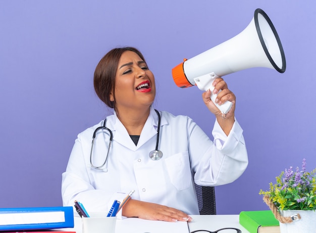 Middle age woman doctor in white coat with stethoscope shouting to megaphone happy and excited sitting at the table over blue background