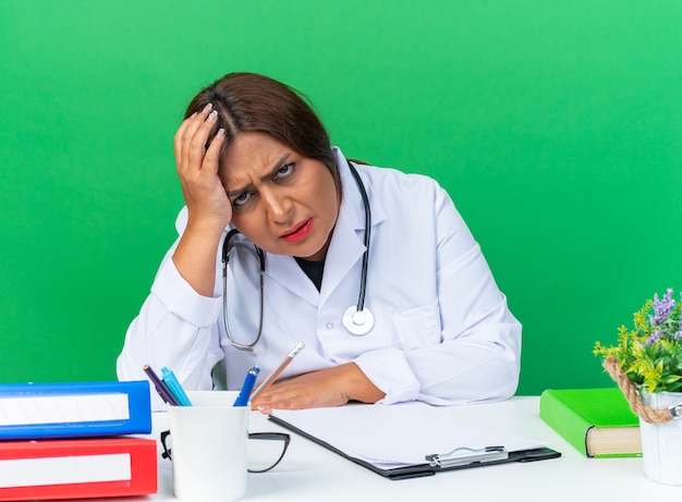 Middle age woman doctor in white coat with stethoscope  tired and bored sitting at the table over green wall