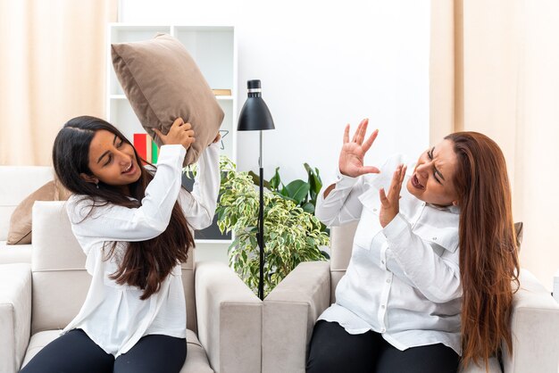 Middle age woman and her young daughter in white shirts and black pants sitting on the chairs quarreling in light living room