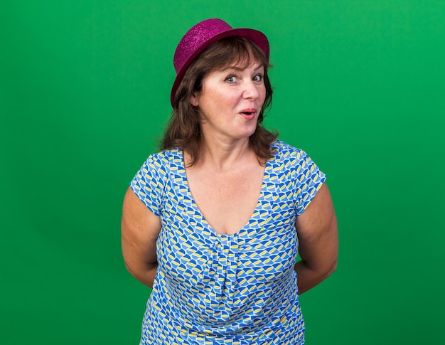 Free photo middle age woman in party hat  happy and surprised celebrating birthday party standing over green wall