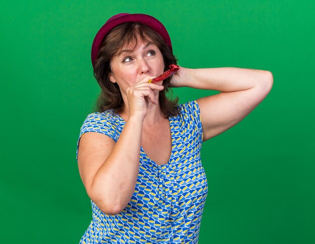 Middle age woman in party hat holding whistle looking up with smile on face celebrating birthday party standing over green wall