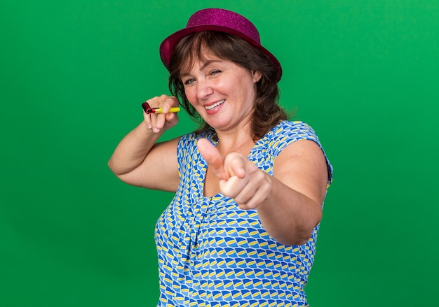 Free Photo middle age woman in party hat holding whistle pointing with index finger  smiling cheerfully celebrating birthday party standing over green wall