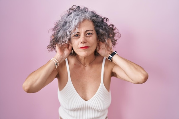 Middle age woman with grey hair standing over pink background trying to hear both hands on ear gesture, curious for gossip. hearing problem, deaf