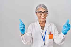 Free photo middle age woman with grey hair wearing scientist robe success sign doing positive gesture with hand thumbs up smiling and happy cheerful expression and winner gesture