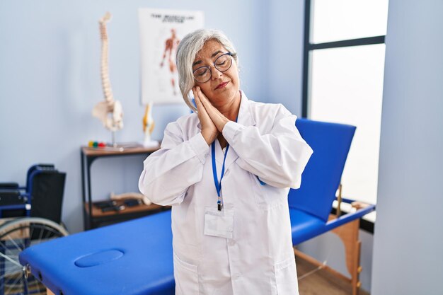 Middle age woman with grey hair working at pain recovery clinic sleeping tired dreaming and posing with hands together while smiling with closed eyes.