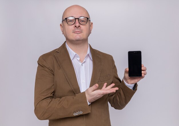 Middle-aged bald man in suit wearing glasses presenting smartphone looking at front smiling confident standing over white wall