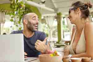 Free photo middle-aged bearded man with cheerful expression pointing thumb at his girlfriend in sunglasses while telling jokes.