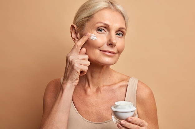 Free photo middle aged beautiful woman applies anti aging cream on face undergoes beauty treatments cares about skin