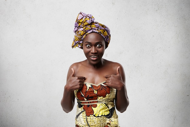 Free photo middle-aged black woman in traditional clothes looking with widely opened eyes having surprised expression pointing at herself while posing against white concrete wall. shocked african model