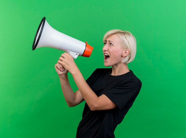 Middle-aged blonde slavic woman standing in profile view shouting in loud speaker looking straight isolated on green background with copy space