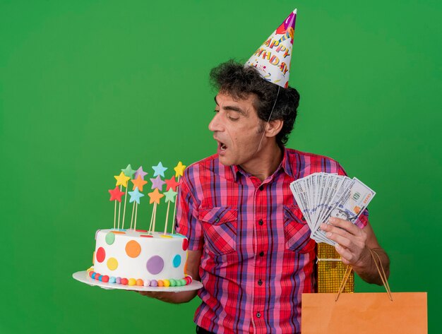 Middle-aged caucasian party man wearing birthday cap holding birthday cake paper bag gift pack and money looking at cake getting ready to bite it isolated on green background