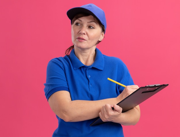 Middle aged delivery woman in blue uniform and cap holding clipboard and pencil looking aside with serious confident expression standing over pink wall