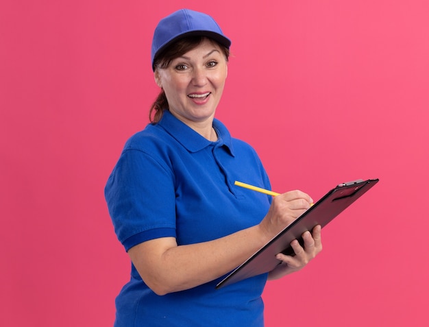 Middle aged delivery woman in blue uniform and cap holding clipboard and pencil writing looking at front hapy and positive smiling cheerfully standing over pink wall