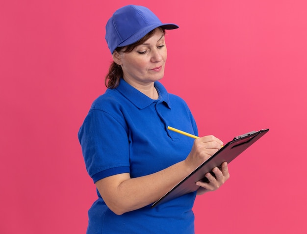 Middle aged delivery woman in blue uniform and cap holding clipboard and pencil writing with serious face standing over pink wall