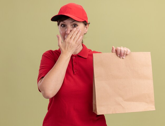 Middle aged delivery woman in red uniform and cap giving paper package looking at front being shocked covering mouth with hand standing over green wall