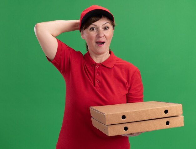 Middle aged delivery woman in red uniform and cap holding pizza boxes looking at front amazed and surprised standing over green wall