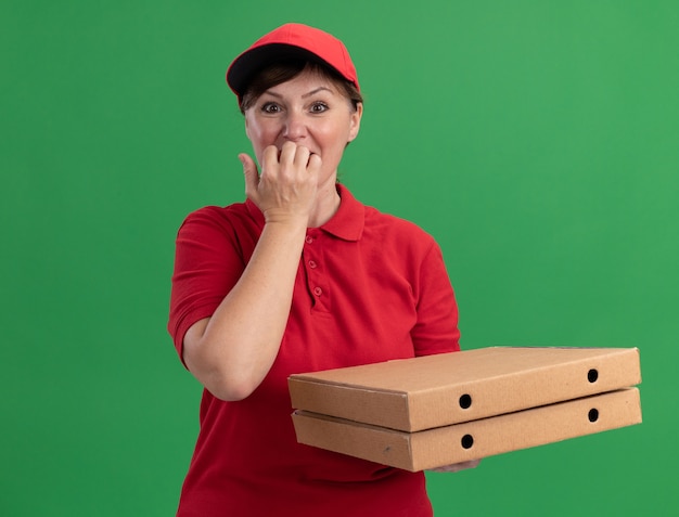 Middle aged delivery woman in red uniform and cap holding pizza boxes looking at front nervous biting nails standing over green wall