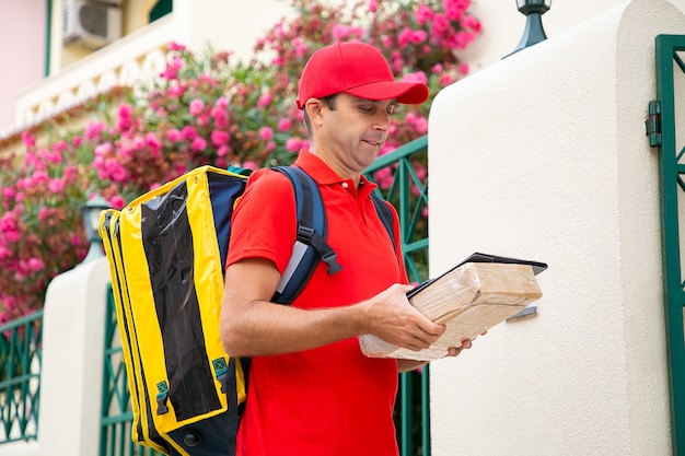 Free photo middle-aged deliveryman holding clipboard and carton box and reading address in receipt. focused postman in red uniform carrying thermo bag and delivering order. home delivery service and post concept