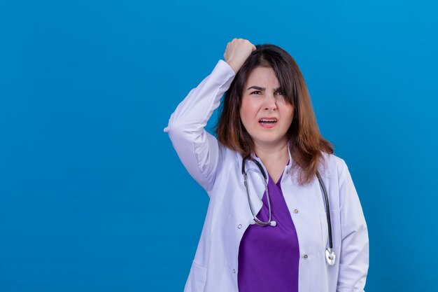 Middle aged doctor wearing white coat and with stethoscope looks confused and disappointed touching her head with hand over blue wall