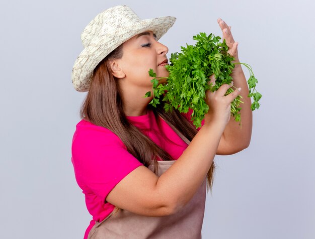 Middle aged gardener woman in apron and hat holding fresh herbs inhaling good aroma standing over white background