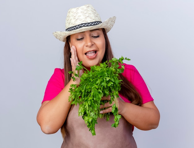 Middle aged gardener woman in apron and hat holding fresh herbs smiling with happy face standing over white background