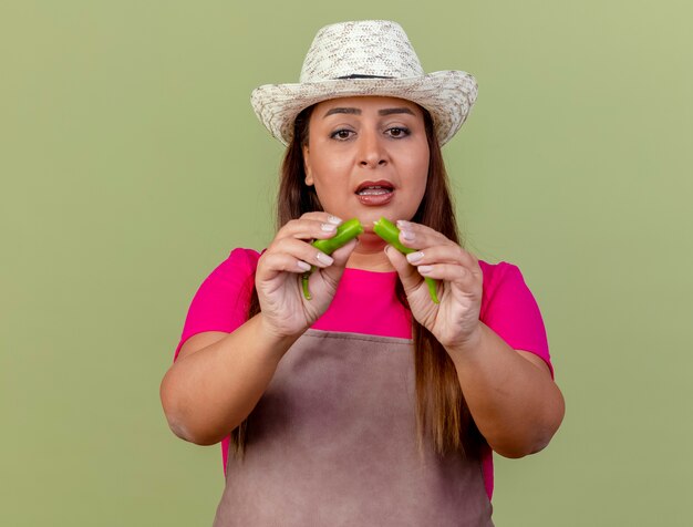 Free photo middle aged gardener woman in apron and hat holding halves of green chili pepper looking confused standing over light background