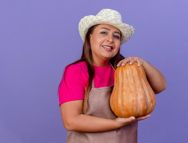 Middle aged gardener woman in apron and hat holding pumpkin smiling with happy face