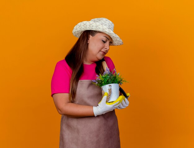 Middle aged gardener woman in apron and hat holding shovel and potted plant looking at plant with smile on face standing over orange background