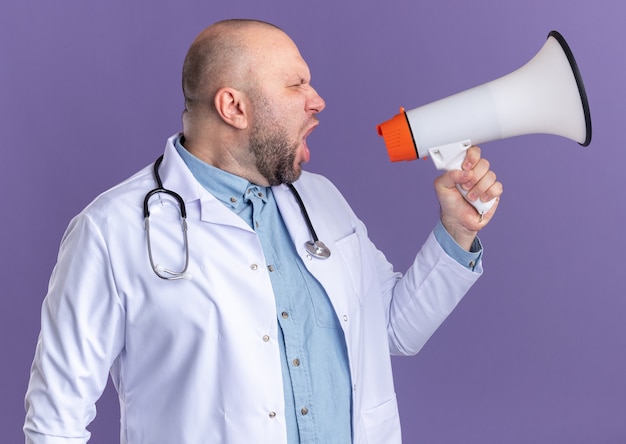 Middle-aged male doctor wearing medical robe and stethoscope looking at side shouting in loud speaker isolated on purple wall