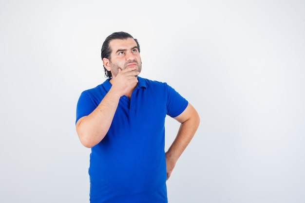 Middle aged man looking up while holding hands on hip and chin in blue t-shirt and looking thoughtful