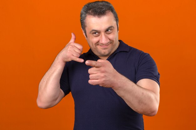 Middle aged man wearing polo shirt smiling doing talking on the telephone gesture and pointing to someone over isolated orange wall