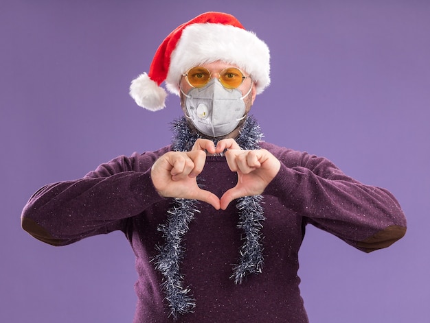 Free Photo middle-aged man wearing santa hat and protective mask with tinsel garland around neck with glasses  doing heart sign isolated on purple wall