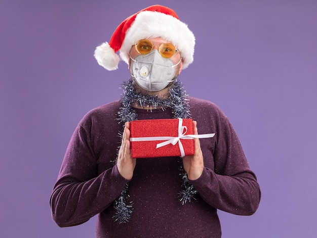 Free Photo middle-aged man wearing santa hat and protective mask with tinsel garland around neck with glasses  holding gift package isolated on purple wall