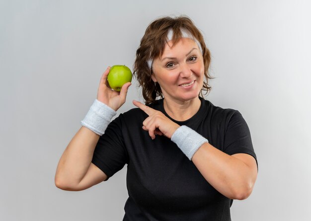 Middle aged sporty woman in black t-shirt with headband holding two green apples pointing with finger to them smiling cheerfully standing over white wall