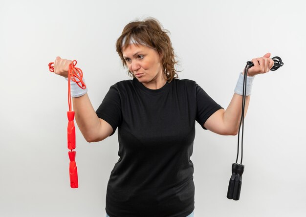 Middle aged sporty woman in black t-shirt with headband holding two skipping ropes looking confused having doubts standing over white wall