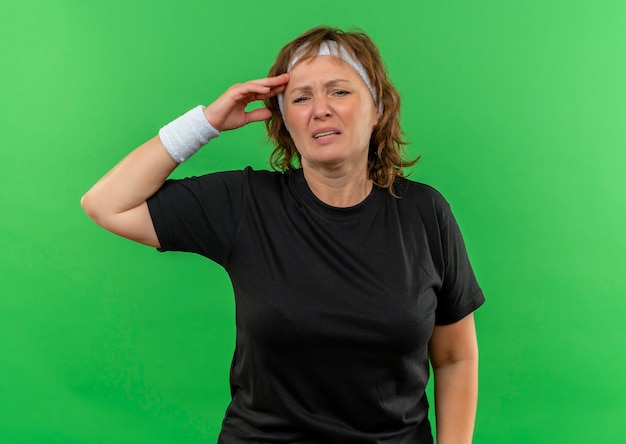 Middle aged sporty woman in black t-shirt with headband looking unwell sick and tired standing over green wall