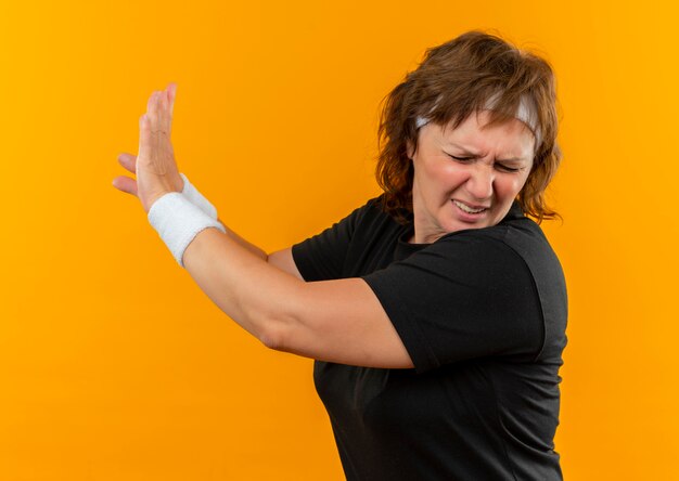 Middle aged sporty woman in black t-shirt with headband making defense gesture with hands standing over orange wall