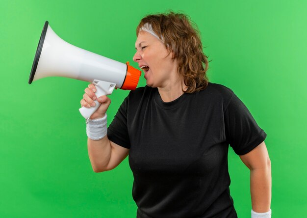 Middle aged sporty woman in black t-shirt with headband shouting to megaphone with aggressive expression standing over green wall