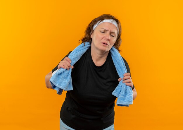 Middle aged sporty woman in black t-shirt with headband and towel on shoulder looking tired and exhausted after workout standing over orange wall