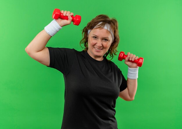 Middle aged sporty woman in black t-shirt with headband working out with two dumbbells looking confident smiling cheerfully standing over green wall