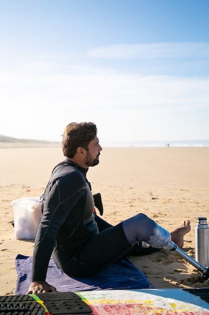 Free photo middle-aged surfer with artificial leg sitting on beach and looking at sea