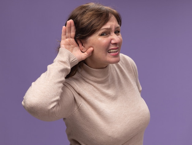 Free Photo middle aged woman in beige turtleneck holding hand over ear trying to listen to gossips standing over purple wall