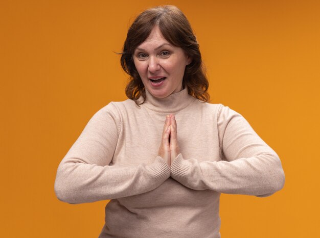 Middle aged woman in beige turtleneck holding hands together like namaste smiling friendly standing over orange wall